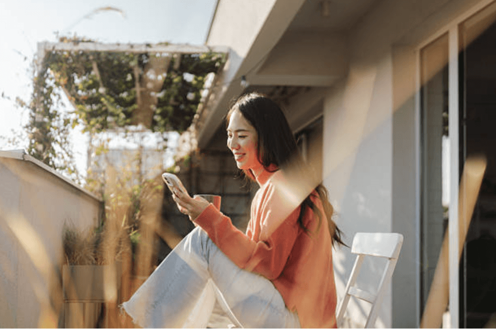 Woman in orange sweater smiles, sitting on balcony chair, using a smartphone.