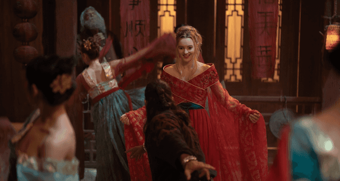Woman in ornate red dress dances joyfully among others in an opulently decorated room.