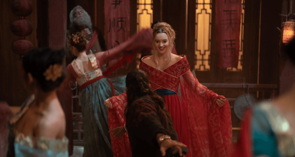 Woman in ornate red dress dances joyfully among others in an opulently decorated room.