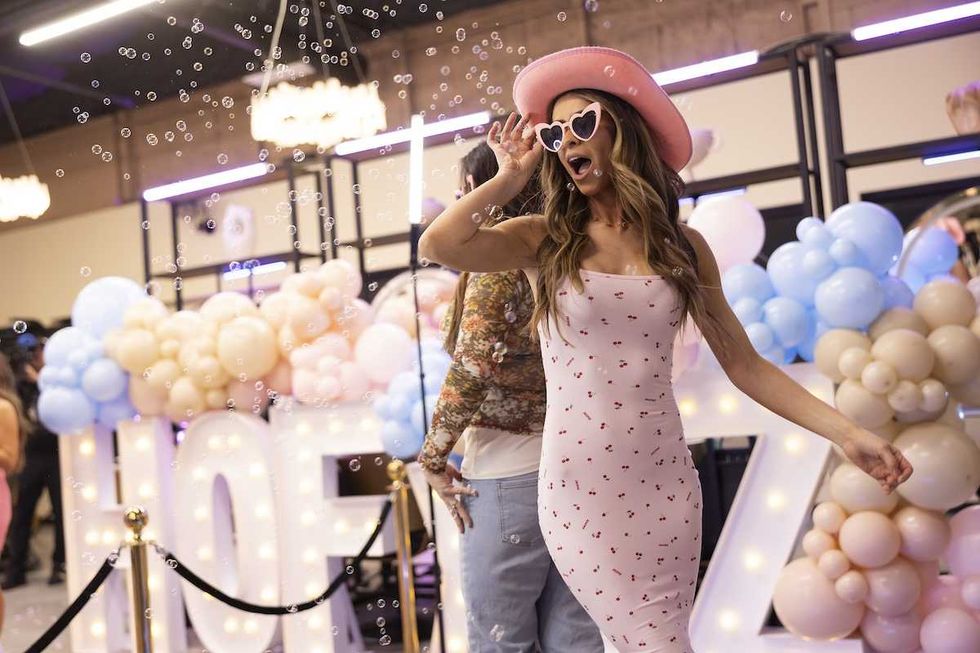 Woman in pink dress and hat poses with bubbles at balloon-decorated event.