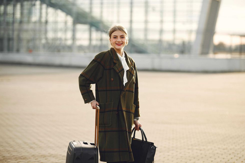 Woman in plaid coat with suitcase and bag outside modern building, smiling.