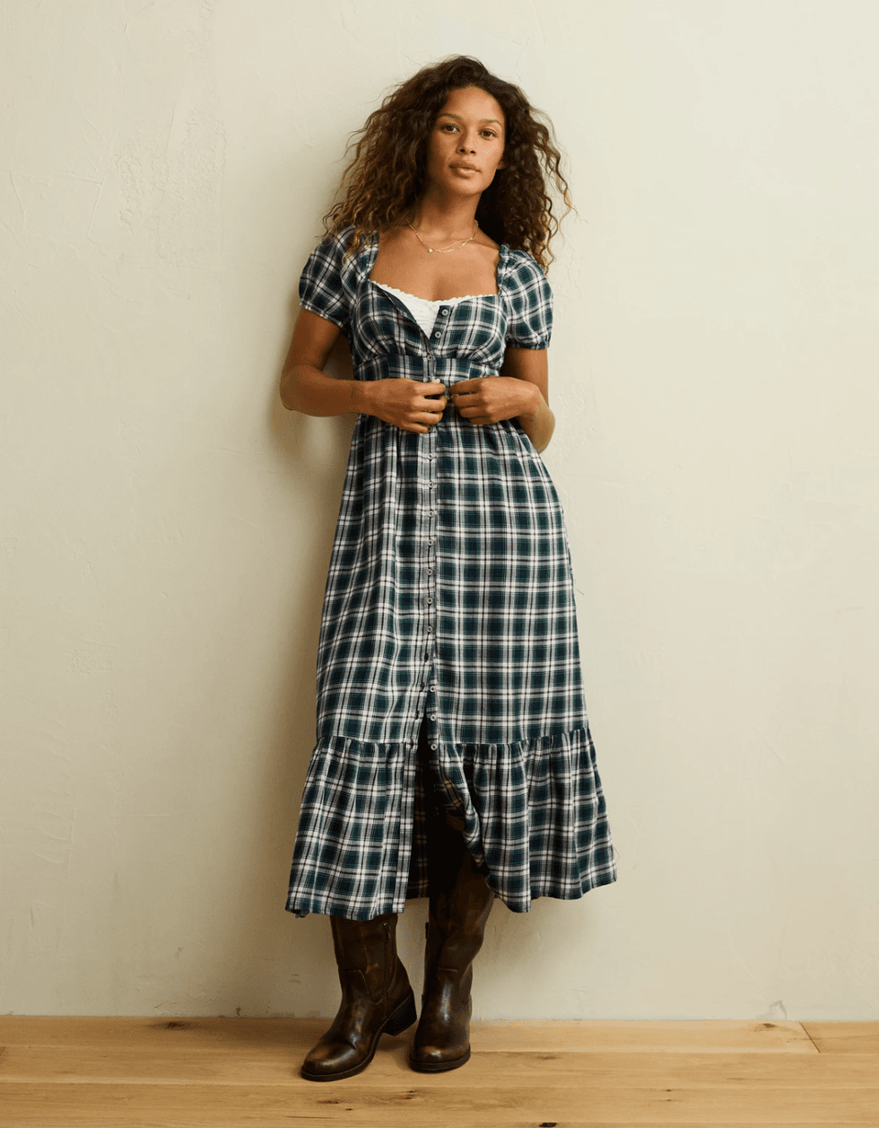 Woman in plaid dress and boots stands against a plain wall.