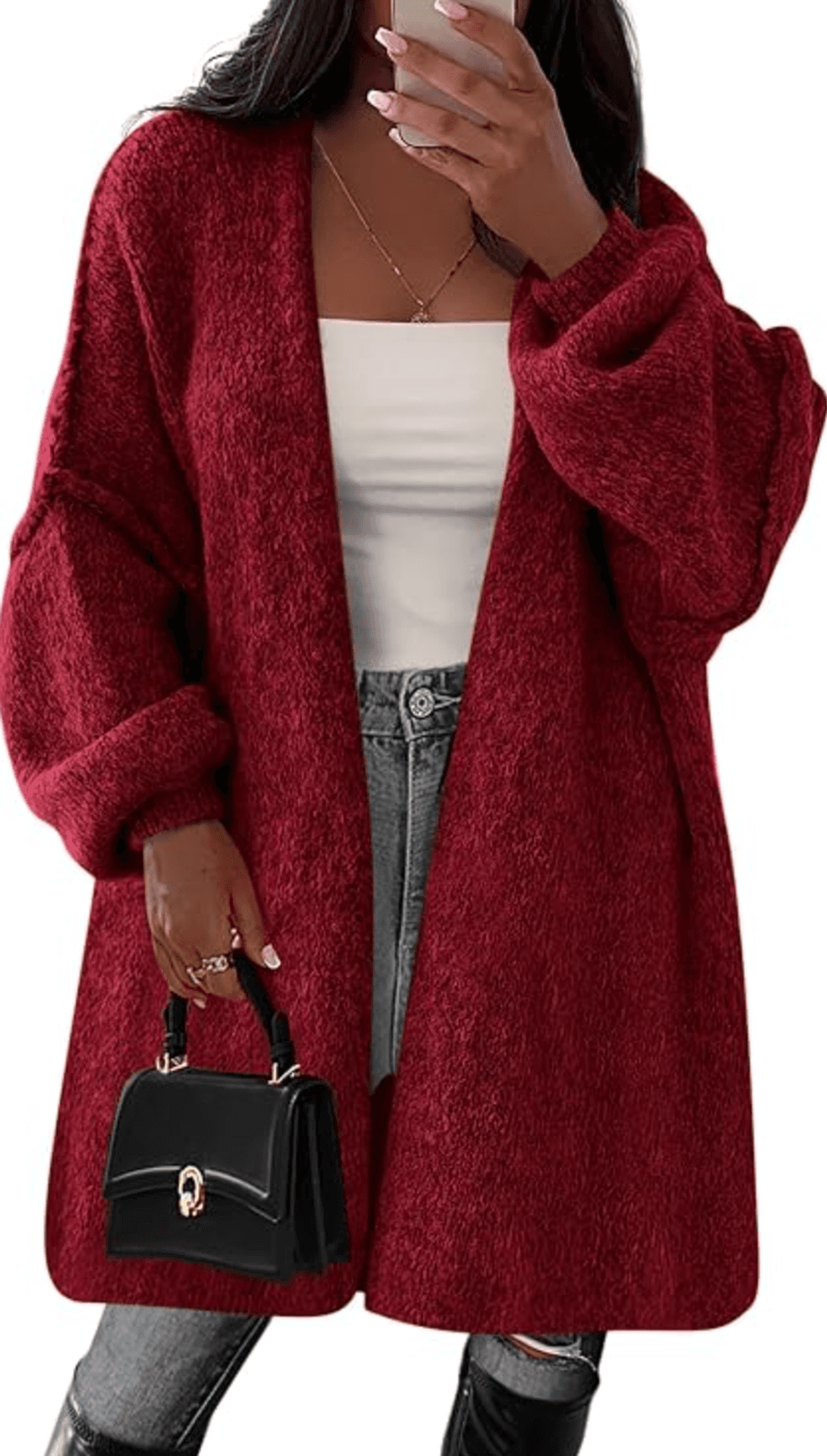 Woman in red cardigan holding phone and black handbag, wearing white top and jeans.