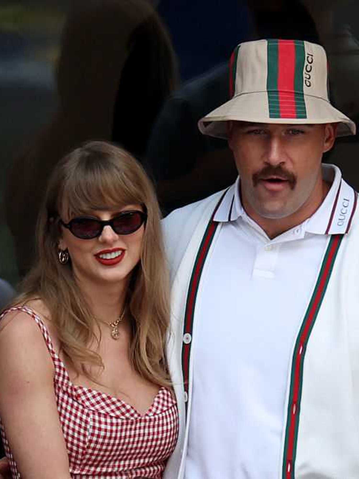 Woman in red dress and sunglasses with man in white outfit and hat at an event.
