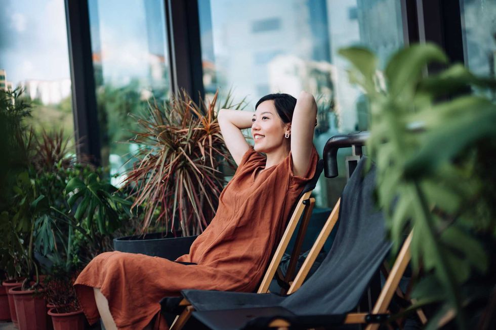 Woman in rust dress relaxing on a balcony chair surrounded by plants.