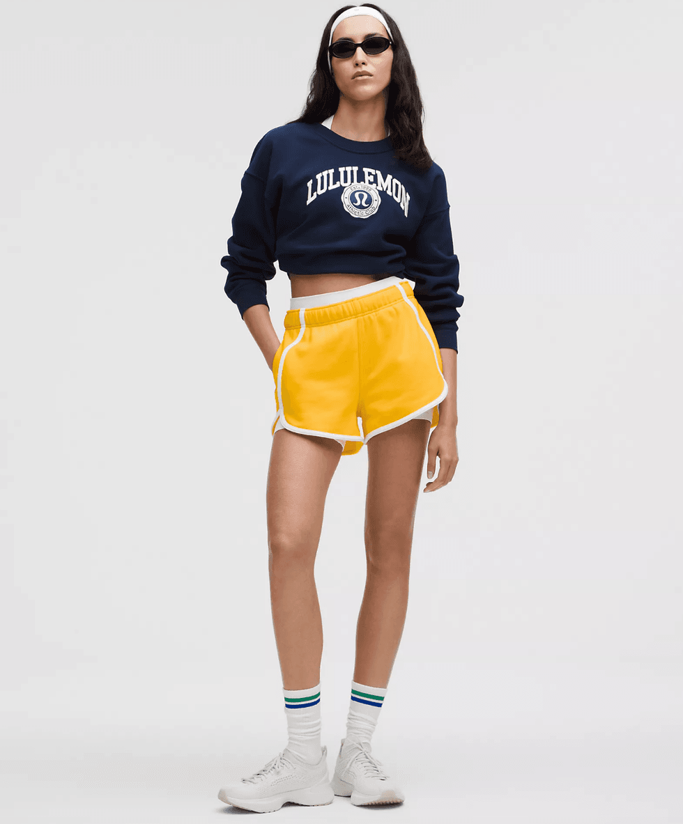 Woman in sporty attire: navy crop sweatshirt, yellow shorts, white shoes, sunglasses, headband.