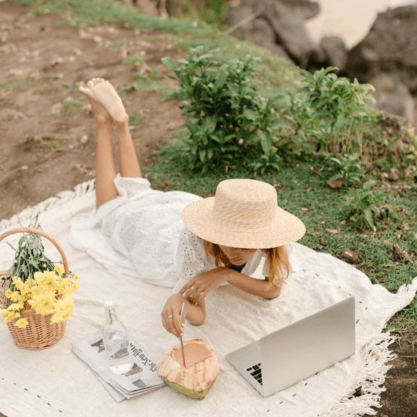 Woman in straw hat working on laptop outdoors with a coconut, flowers, and a bottle nearby.