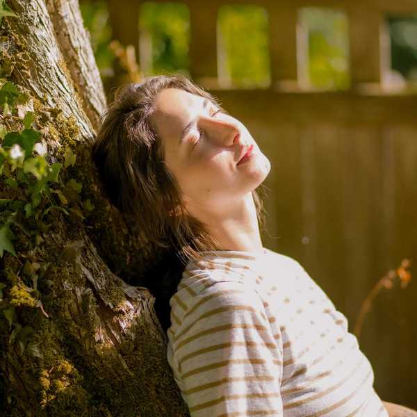 Woman in striped shirt leans against a tree, eyes closed, enjoying the sunlight.