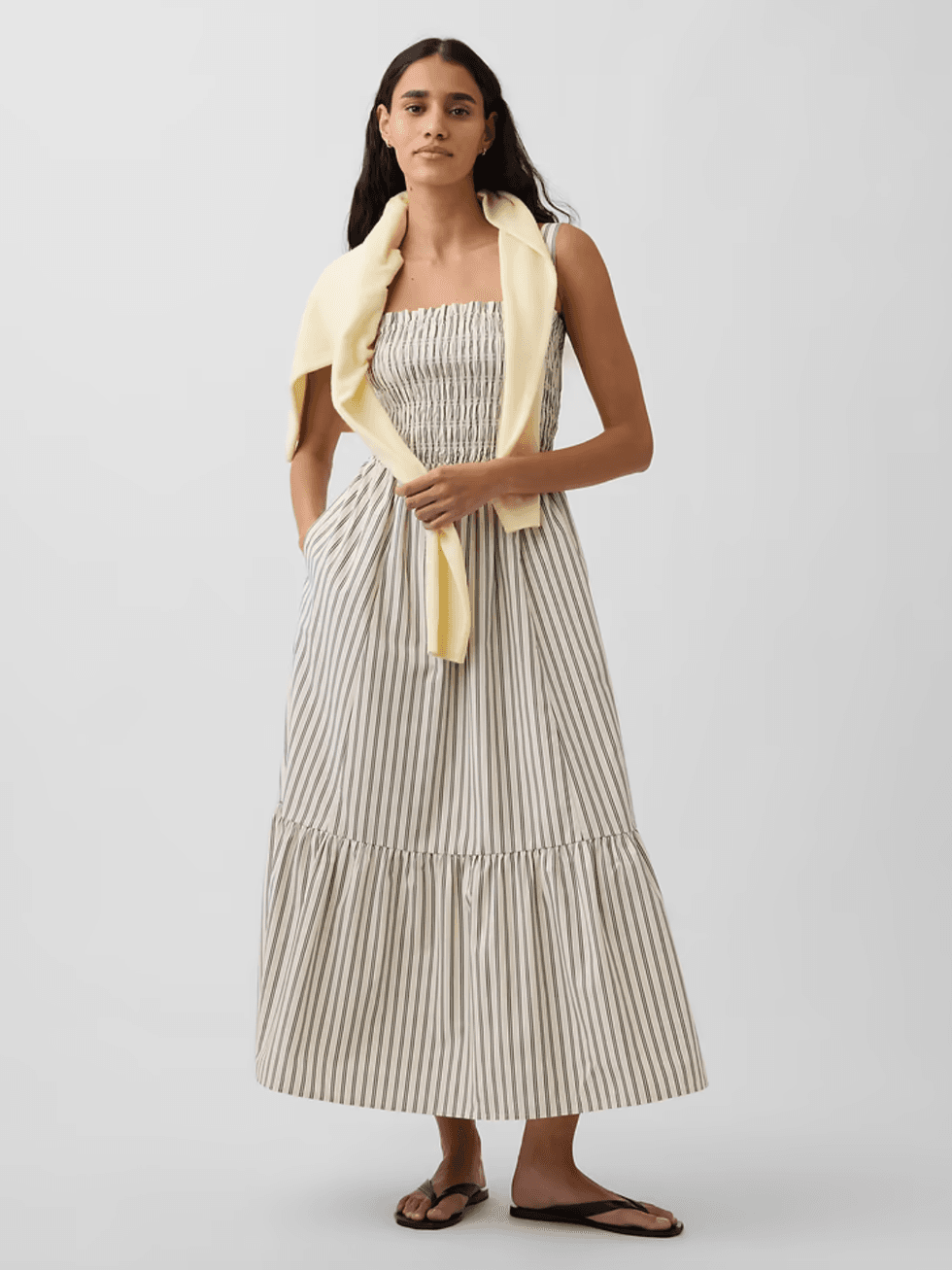 Woman in striped sundress with yellow sweater over shoulders, wearing sandals.