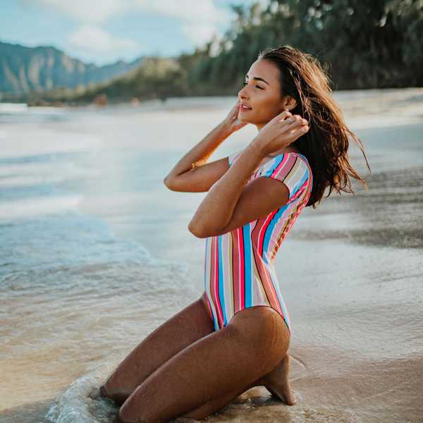 Woman in striped swimsuit kneels on beach, touching hair, with mountains and trees in background.