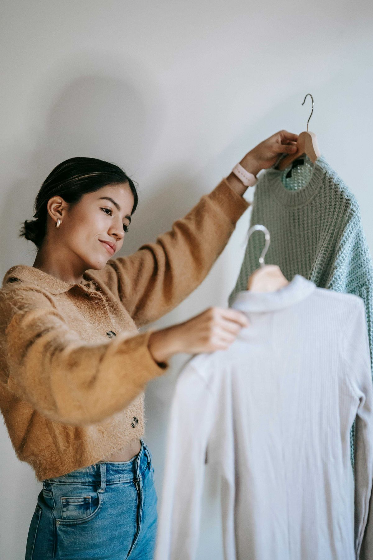 Woman in sweater holding two garments on hangers, deciding between them.