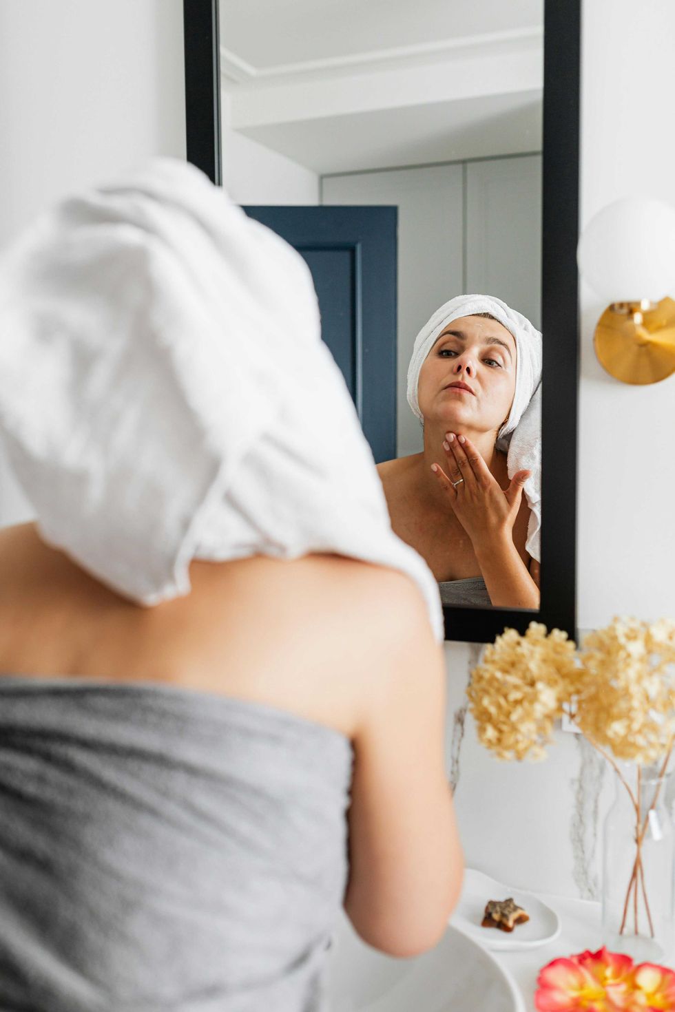 Woman in towel, inspecting face in mirror, bathroom setting.