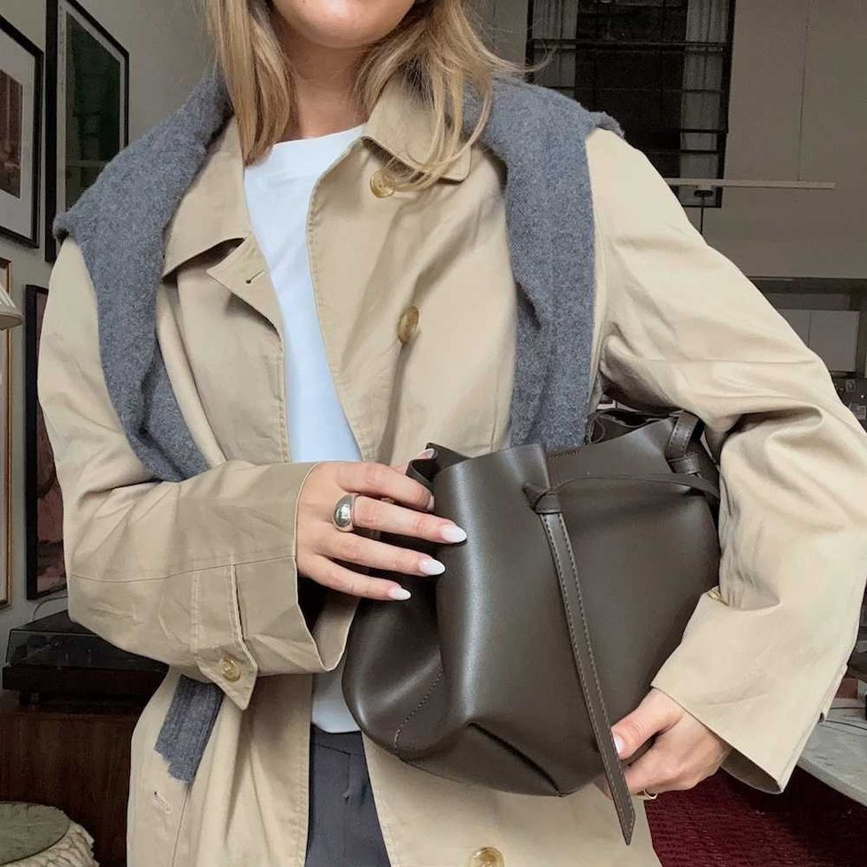 Woman in trench coat holding a dark leather bag indoors.