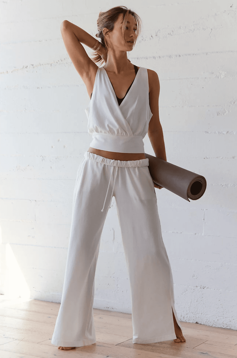 Woman in white activewear holding a yoga mat, standing by a white wall.