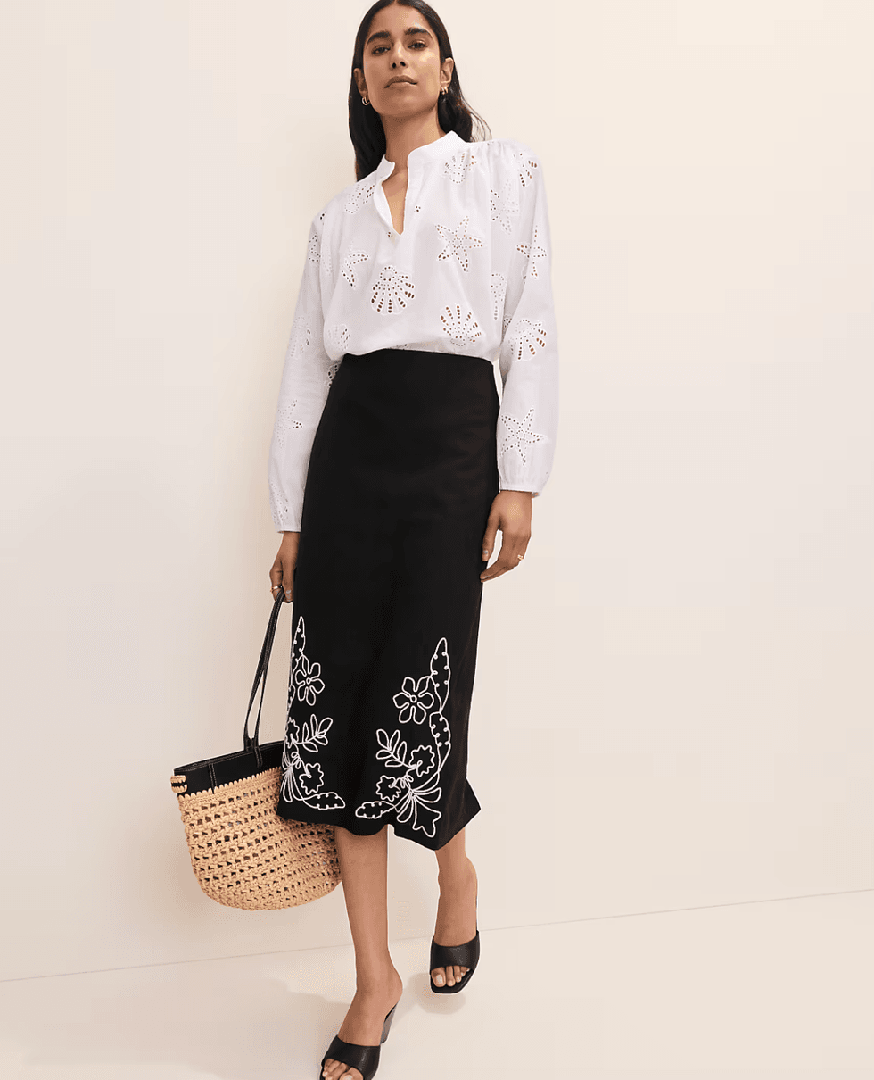 Woman in white blouse and black skirt with floral designs, holding a woven bag.