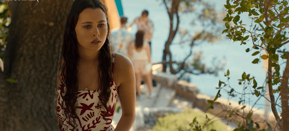 Woman in white floral dress stands thoughtfully by seaside, surrounded by trees.
