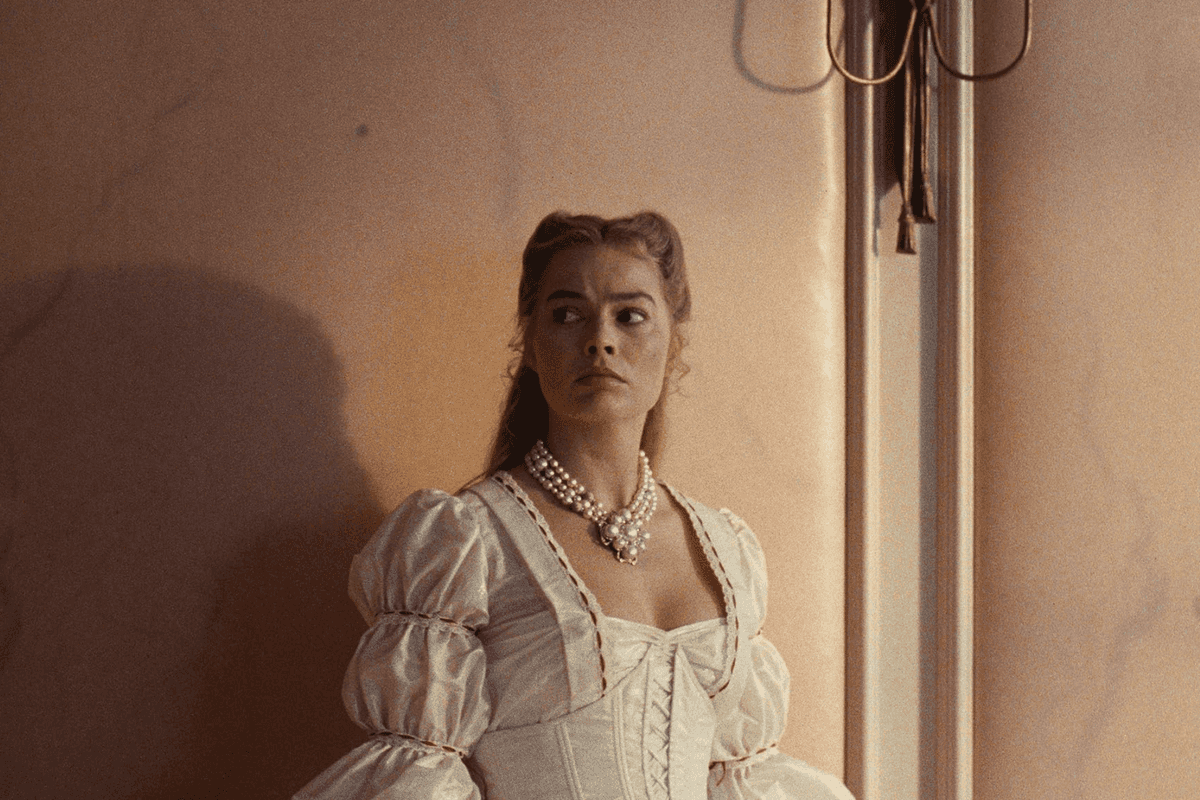 Woman in white gown and pearls stands by a wall, looking to the side.