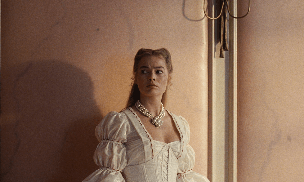 Woman in white gown and pearls stands by a wall, looking to the side.
