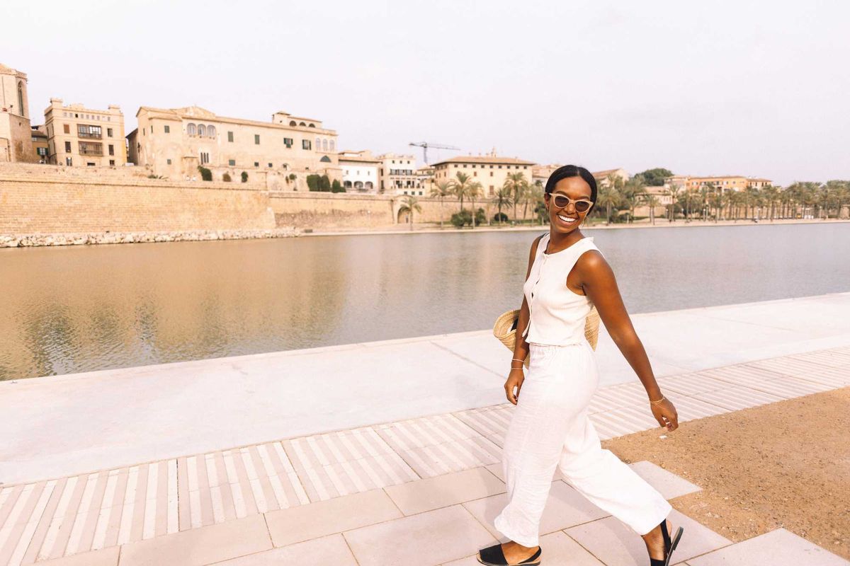 Woman in white jumpsuit smiling by a waterfront with buildings in the background.