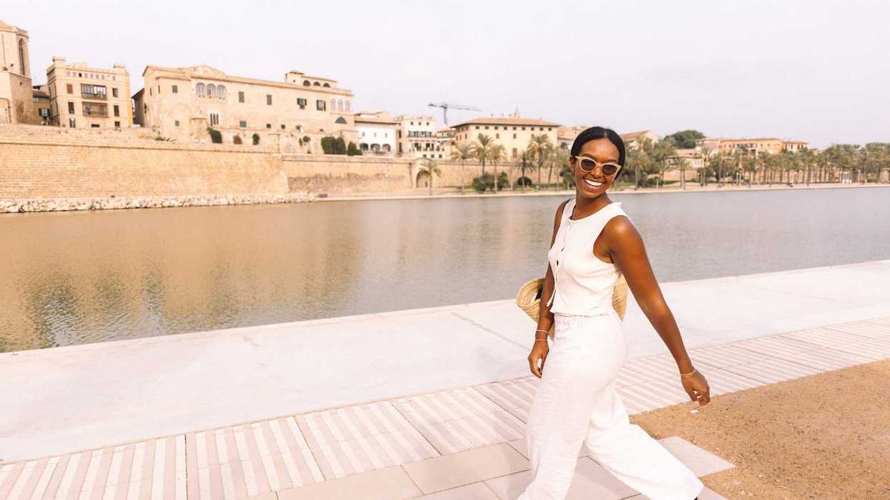 Woman in white jumpsuit smiling by a waterfront with buildings in the background.