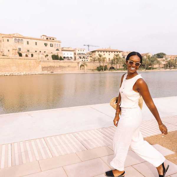 Woman in white jumpsuit smiling by a waterfront with buildings in the background.