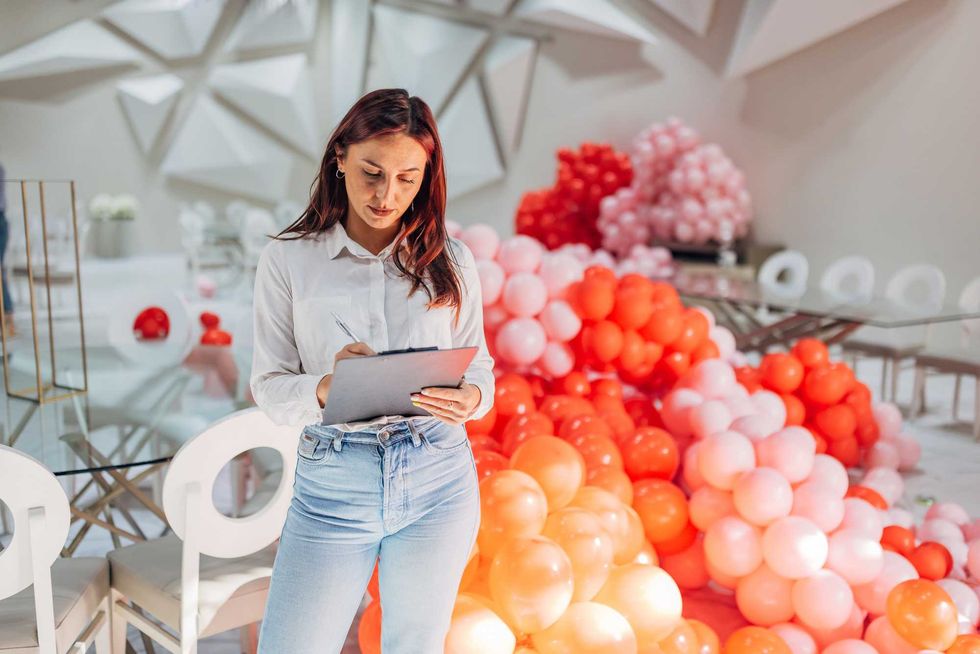 Woman in white shirt and jeans writing on clipboard, surrounded by pink and orange balloons.