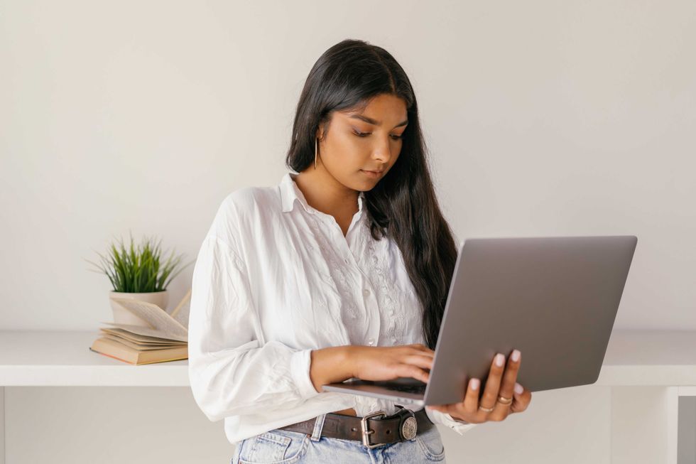 Woman in white shirt typing on a laptop, standing near books and plant.