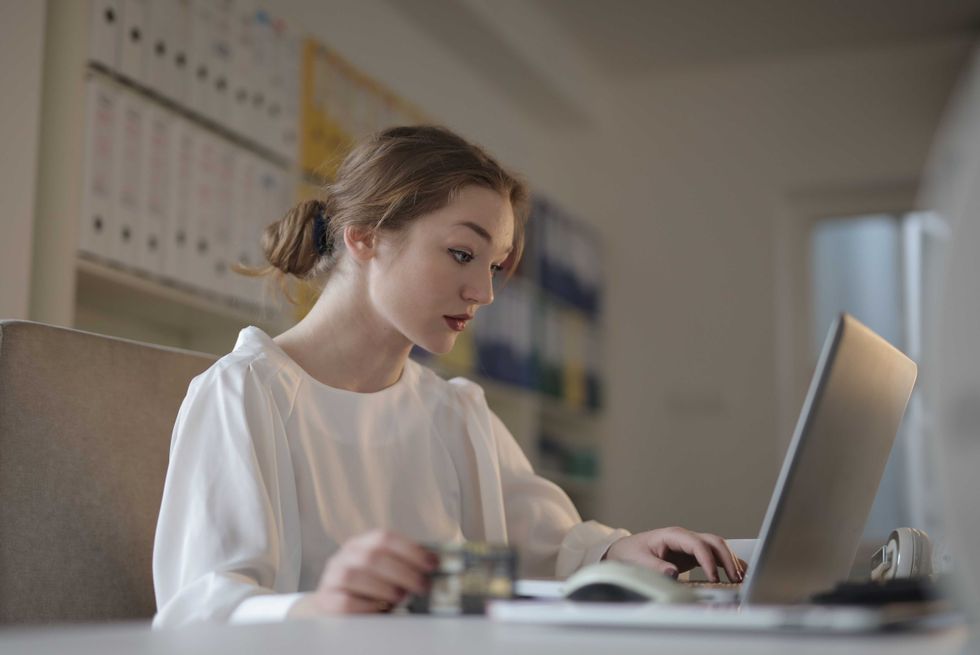 Woman in white shirt working on a laptop at a desk in an office setting.