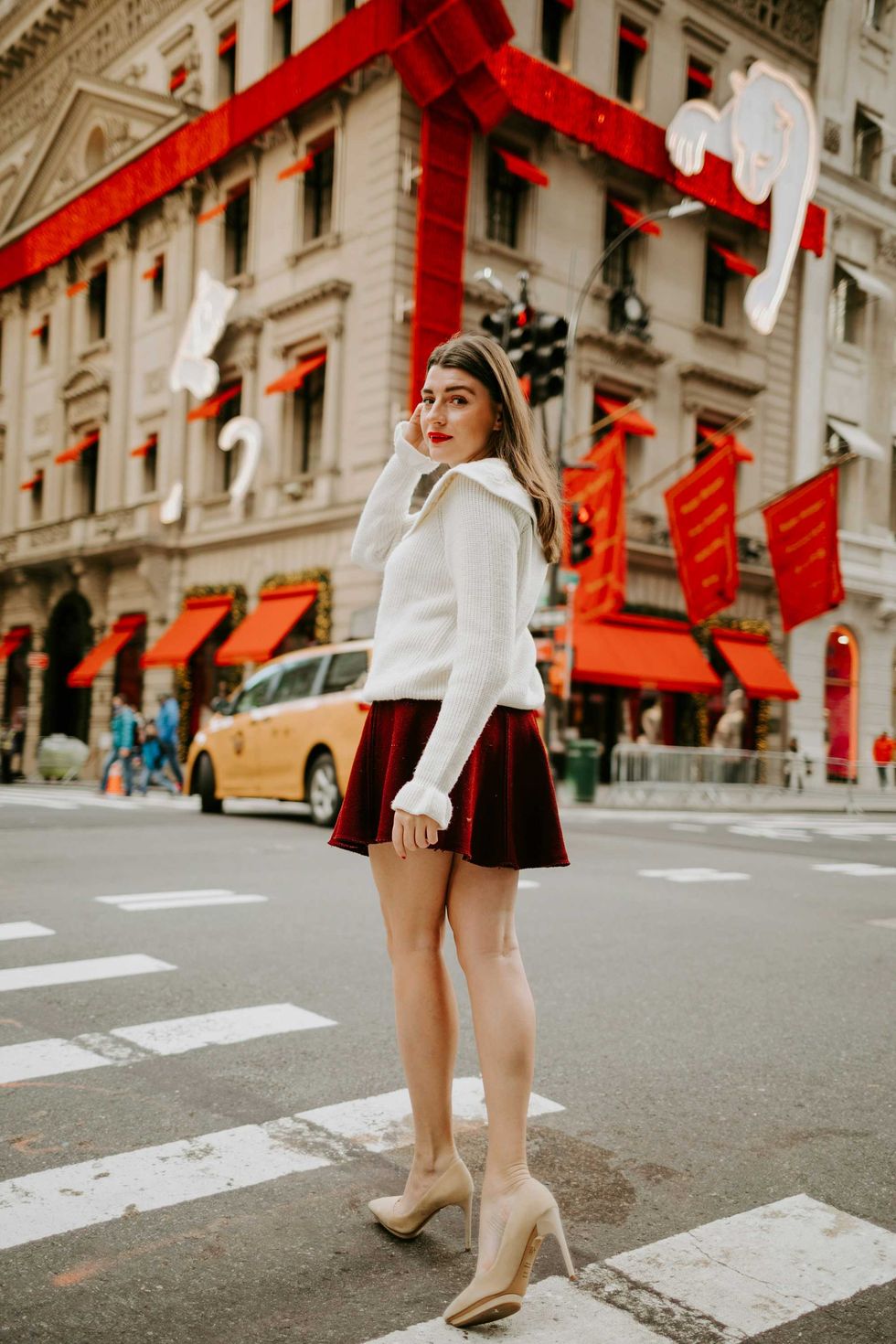 Woman in white sweater and red skirt poses on city street, festive building in background.