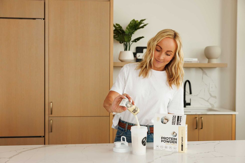 Woman in white t-shirt pouring protein powder into a shaker in a modern kitchen.