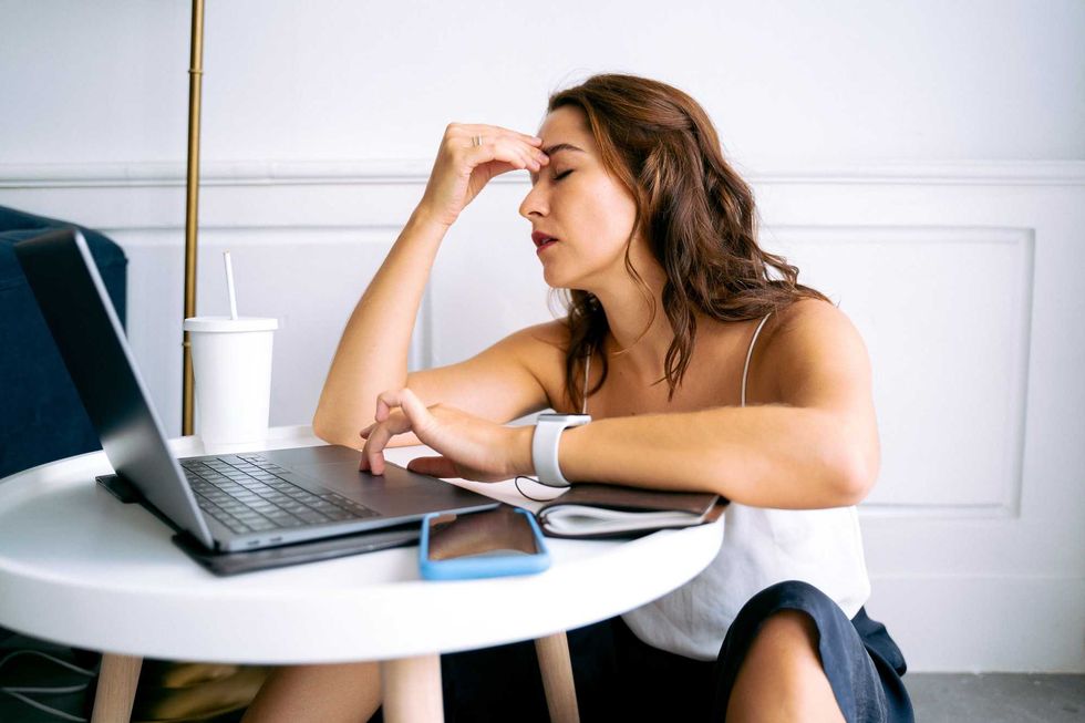 Woman in white tank top, stressed at laptop on round table, holding head with hand.