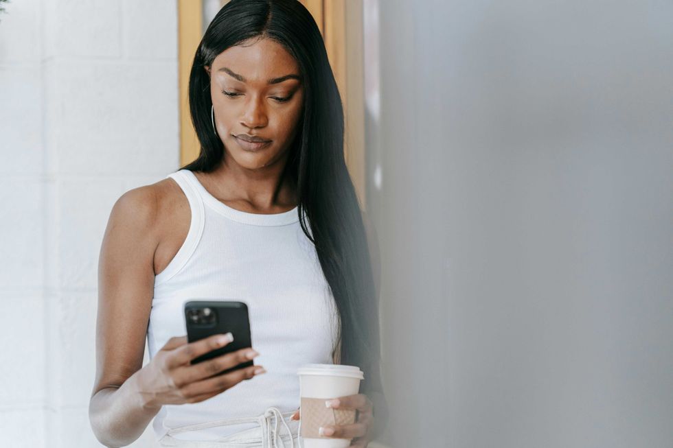 Woman in white tank top texting on phone, holding a coffee cup.