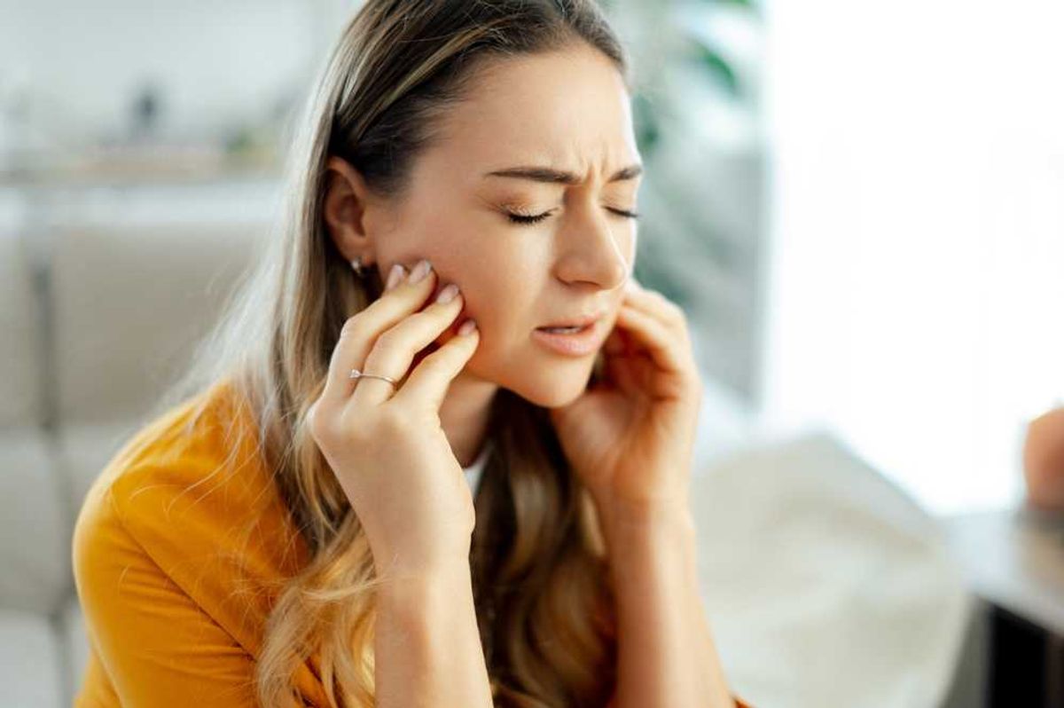 Woman in yellow shirt experiencing jaw pain, eyes closed, hands on cheeks.