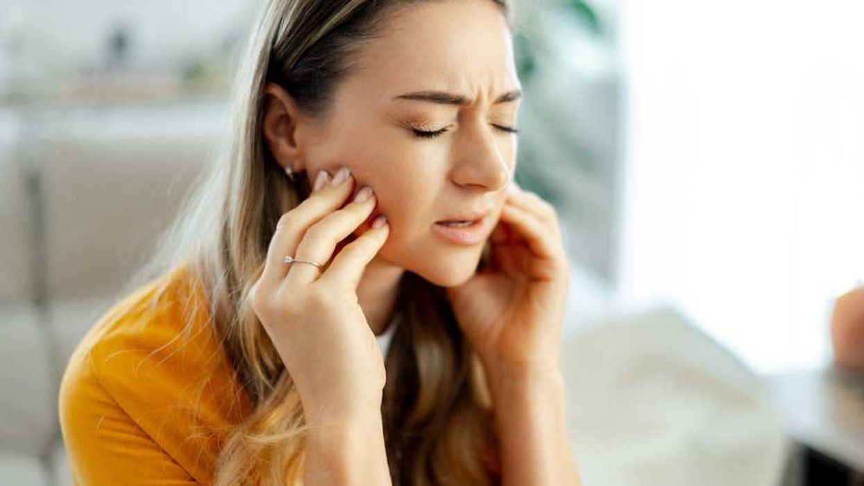Woman in yellow shirt experiencing jaw pain, eyes closed, hands on cheeks.