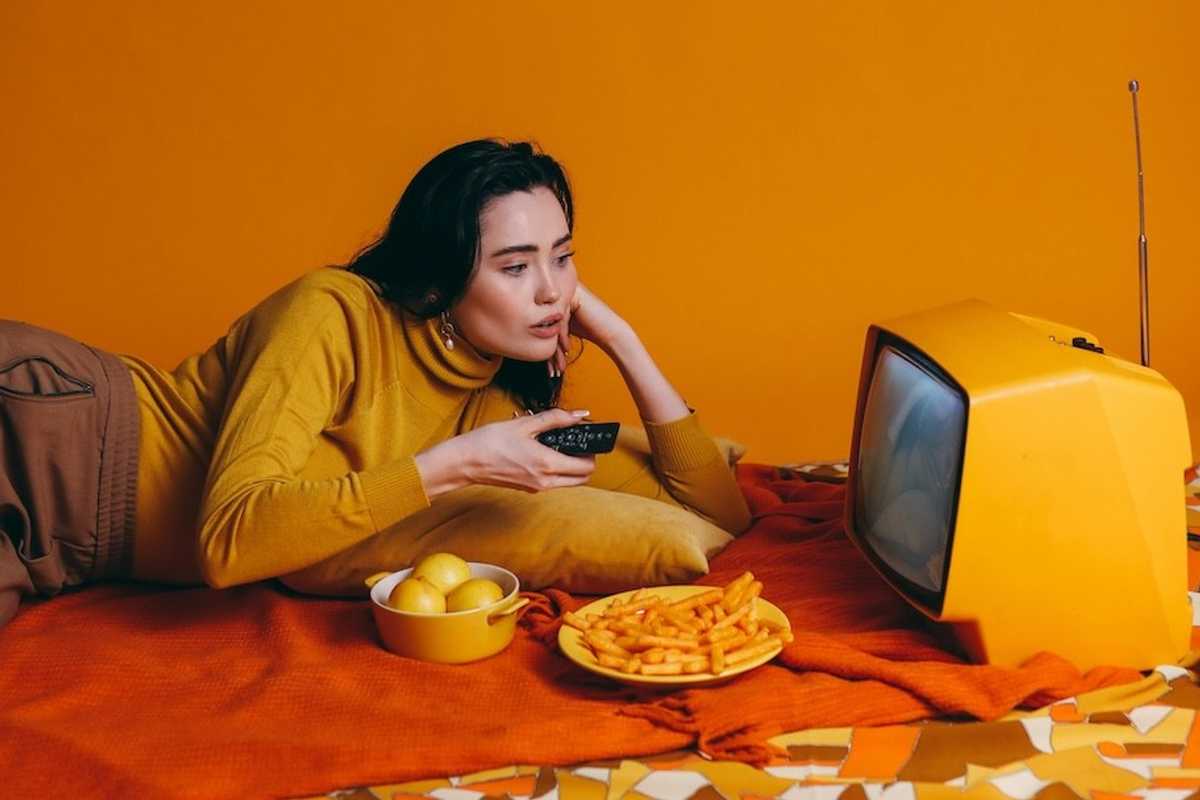 Woman in yellow watching TV with snacks on an orange-themed setup.
