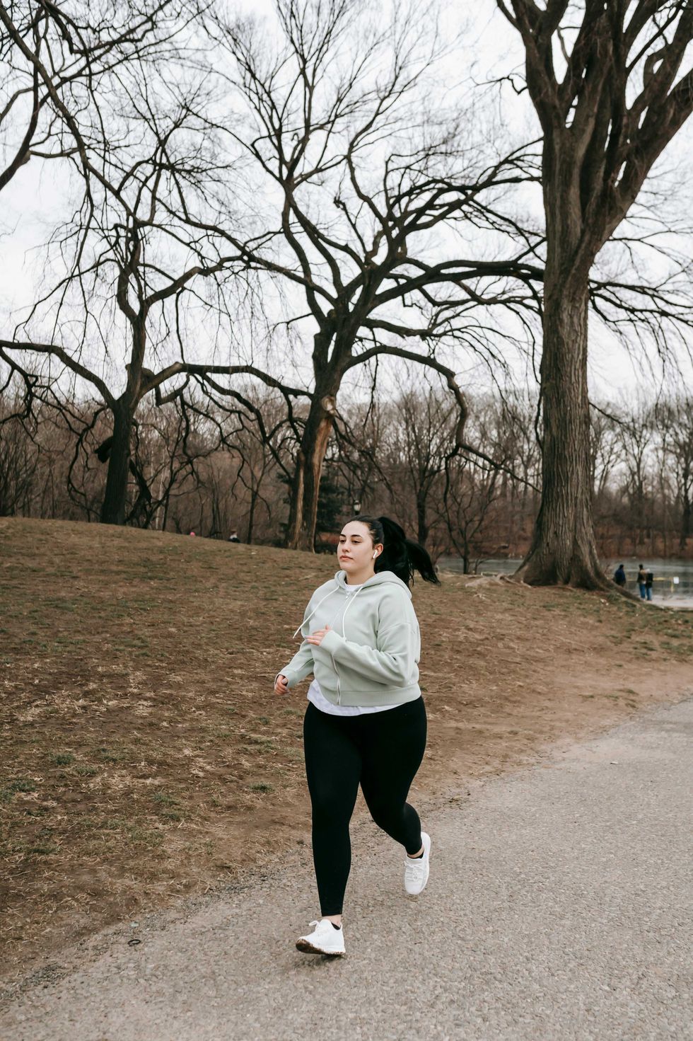 Woman jogging on park path with bare trees in background.