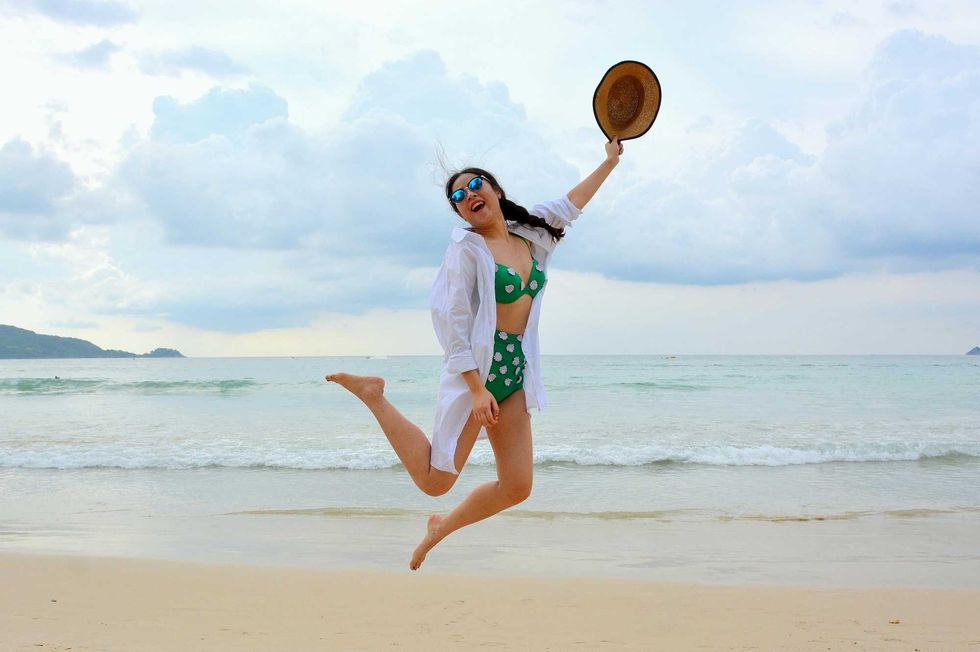Woman jumping joyfully on a beach in a green swimsuit, holding a hat, overcast sky.