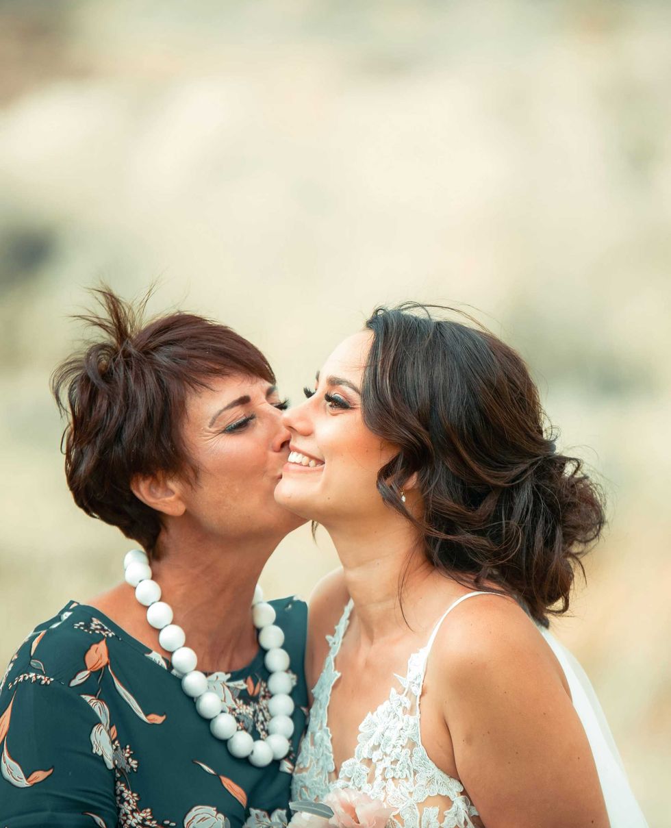 Woman kisses a smiling bride on the cheek outdoors.
