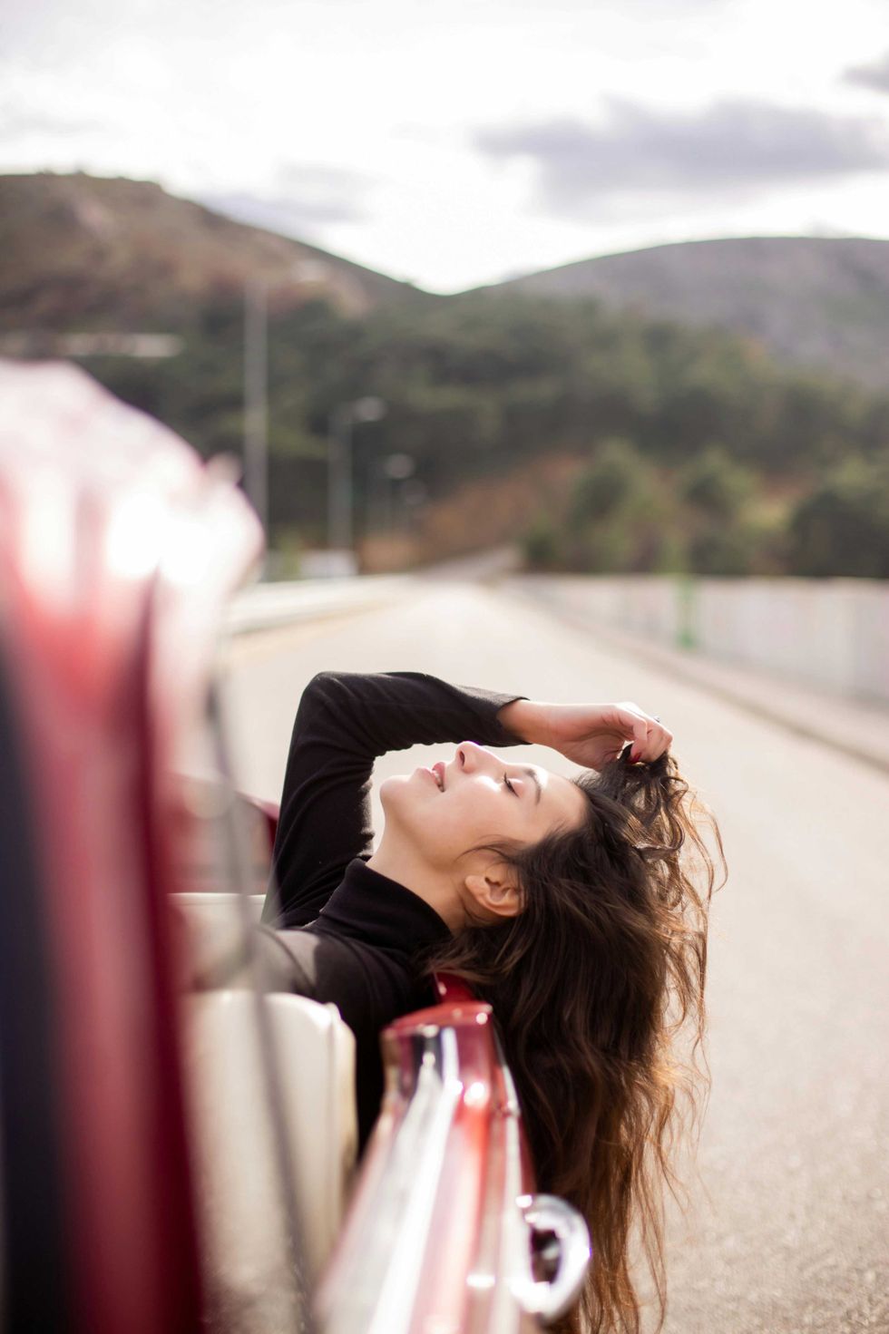Woman leaning out of a red car, enjoying the breeze on a sunny road.