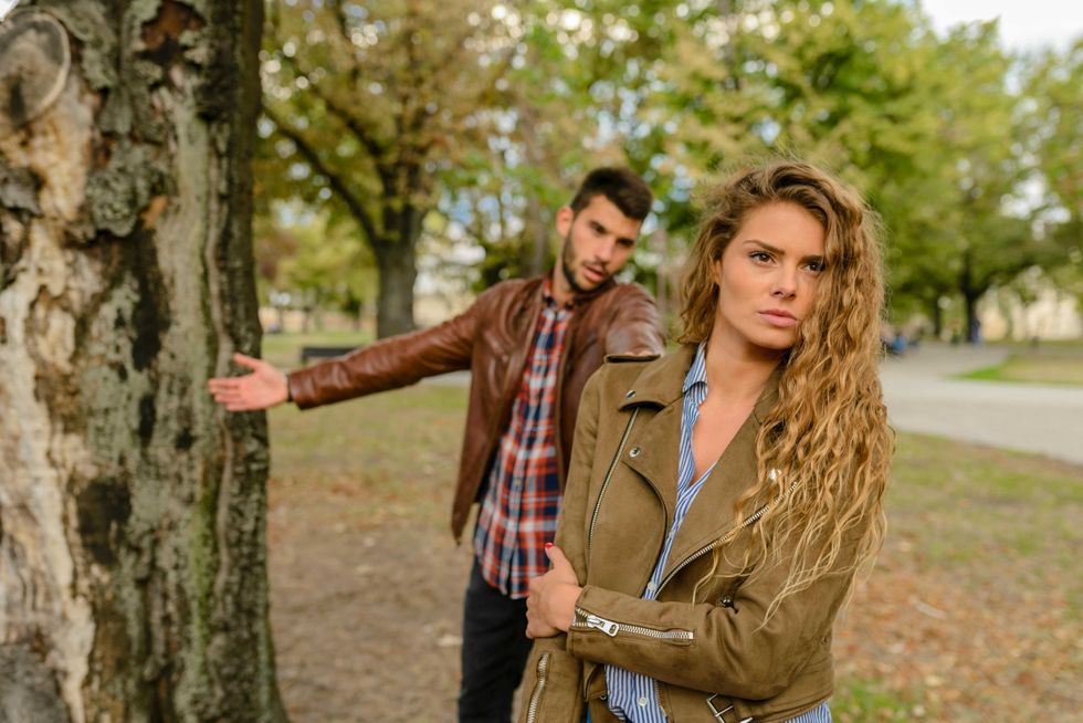 Woman looking serious, man gesturing behind her, both in a park with trees.