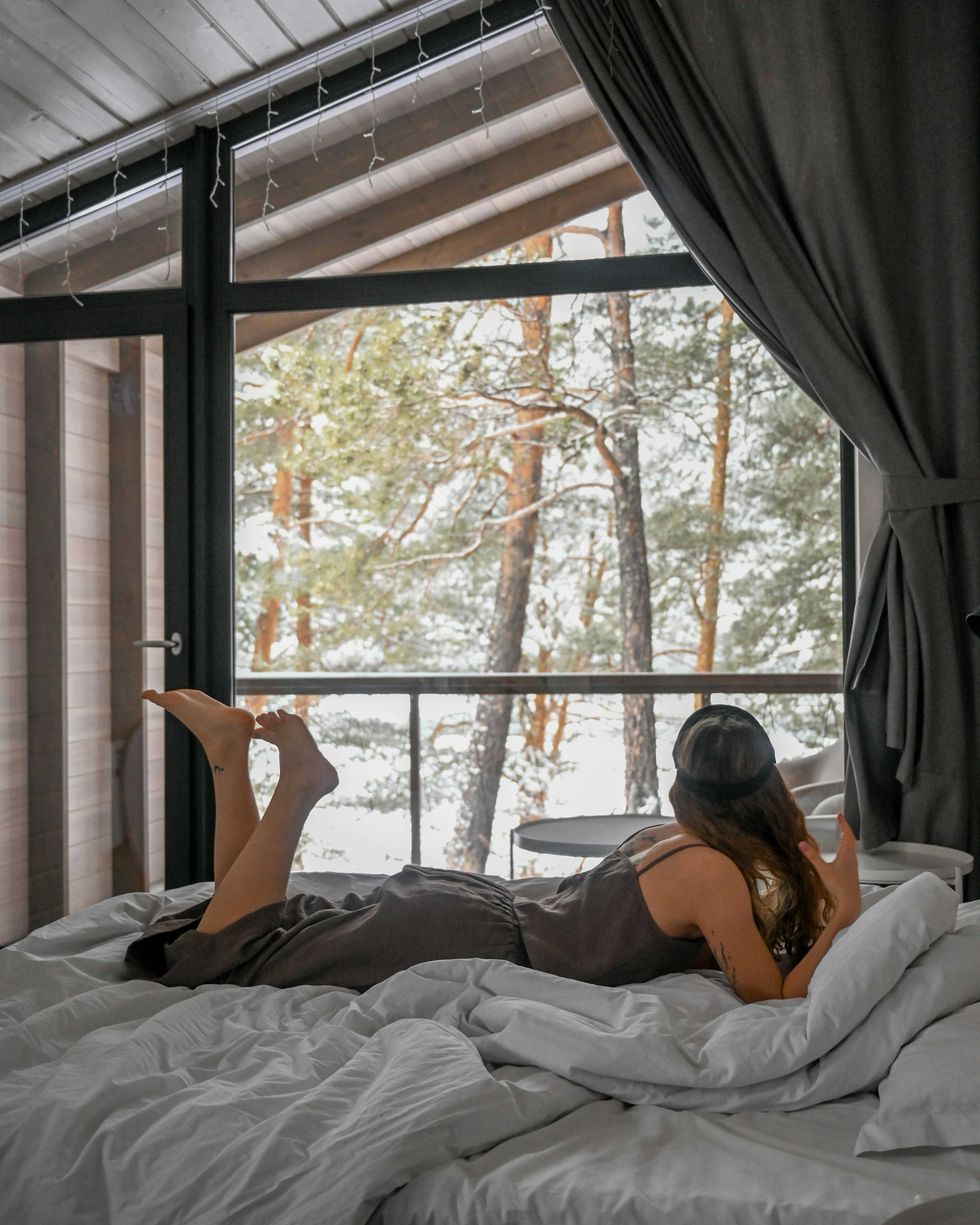 Woman lounging on bed, gazing out at snowy forest through large windows.