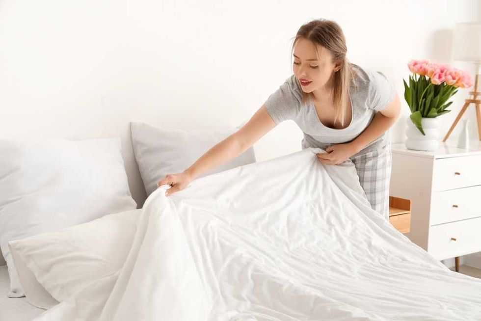 Woman making a bed in a bright room with flowers on a nightstand.
