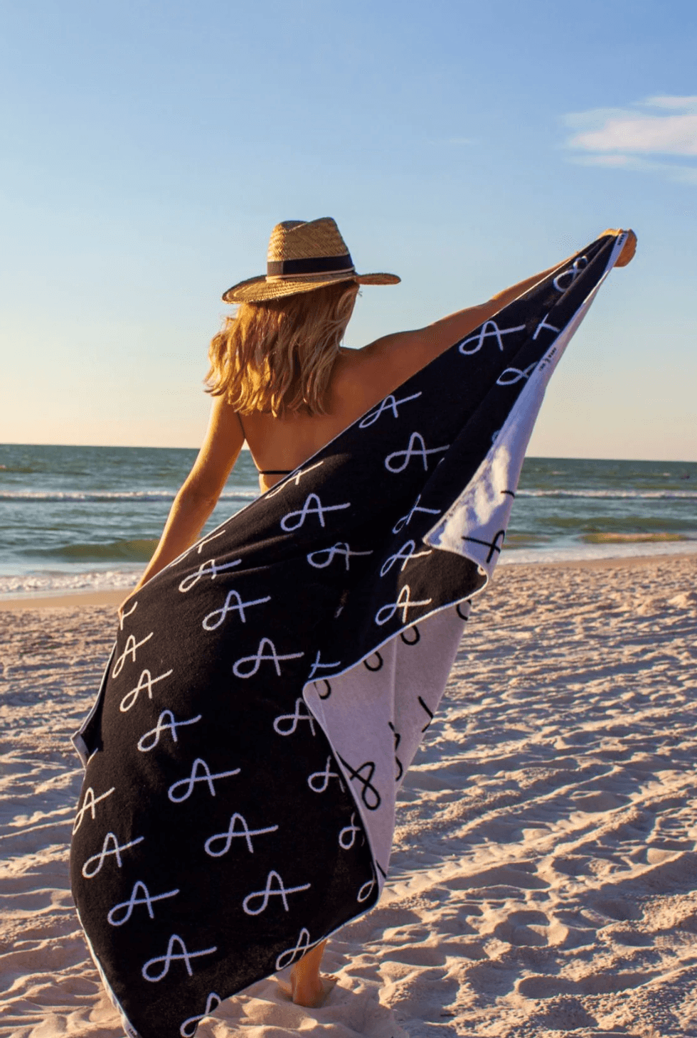 Woman on beach in straw hat holding patterned towel with letter "A" design.