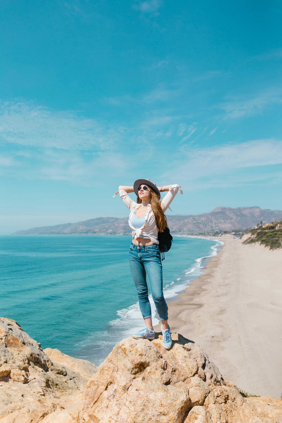 Woman on cliff edge, arms stretched, with ocean and clear sky in background.