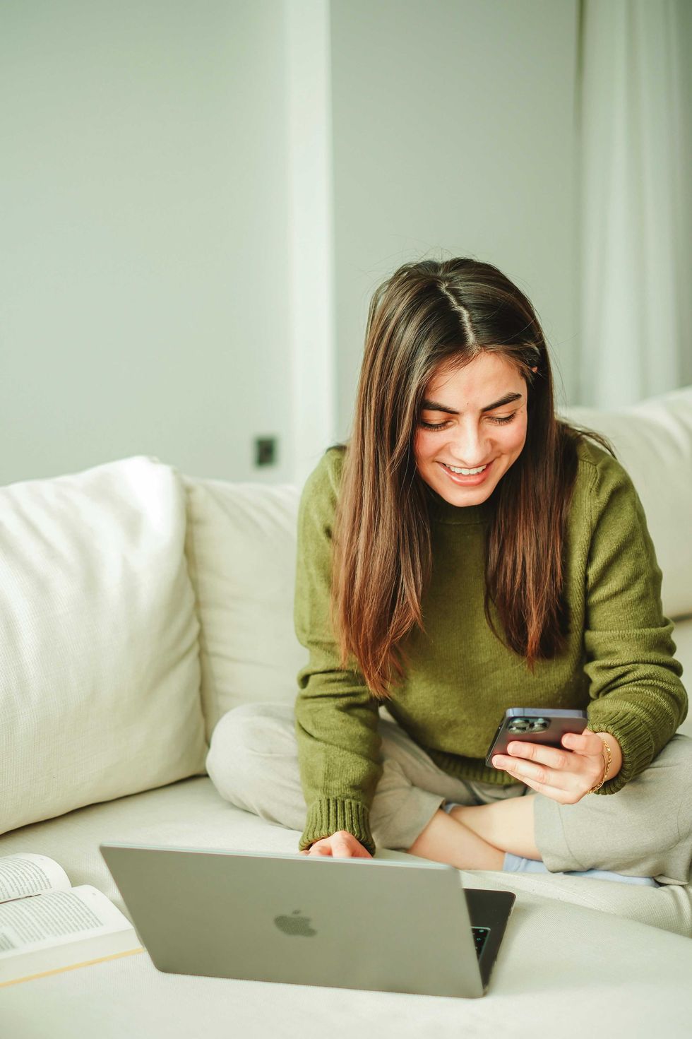 Woman on couch, using laptop and smartphone, smiling.