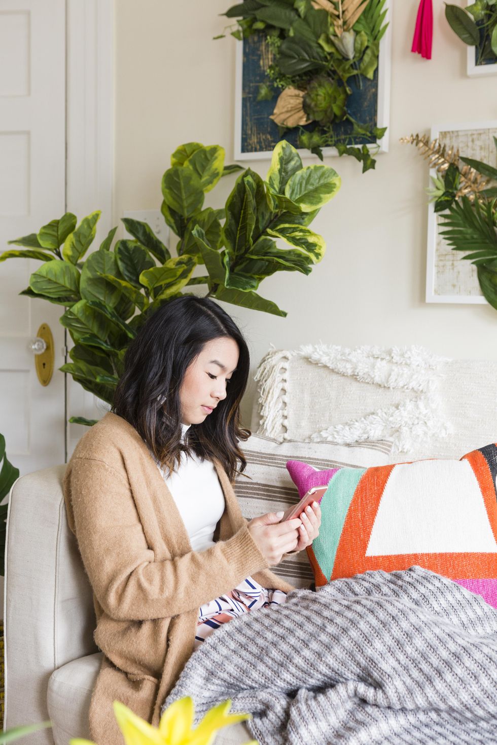 Woman on couch using phone, surrounded by colorful pillows and leafy decor.
