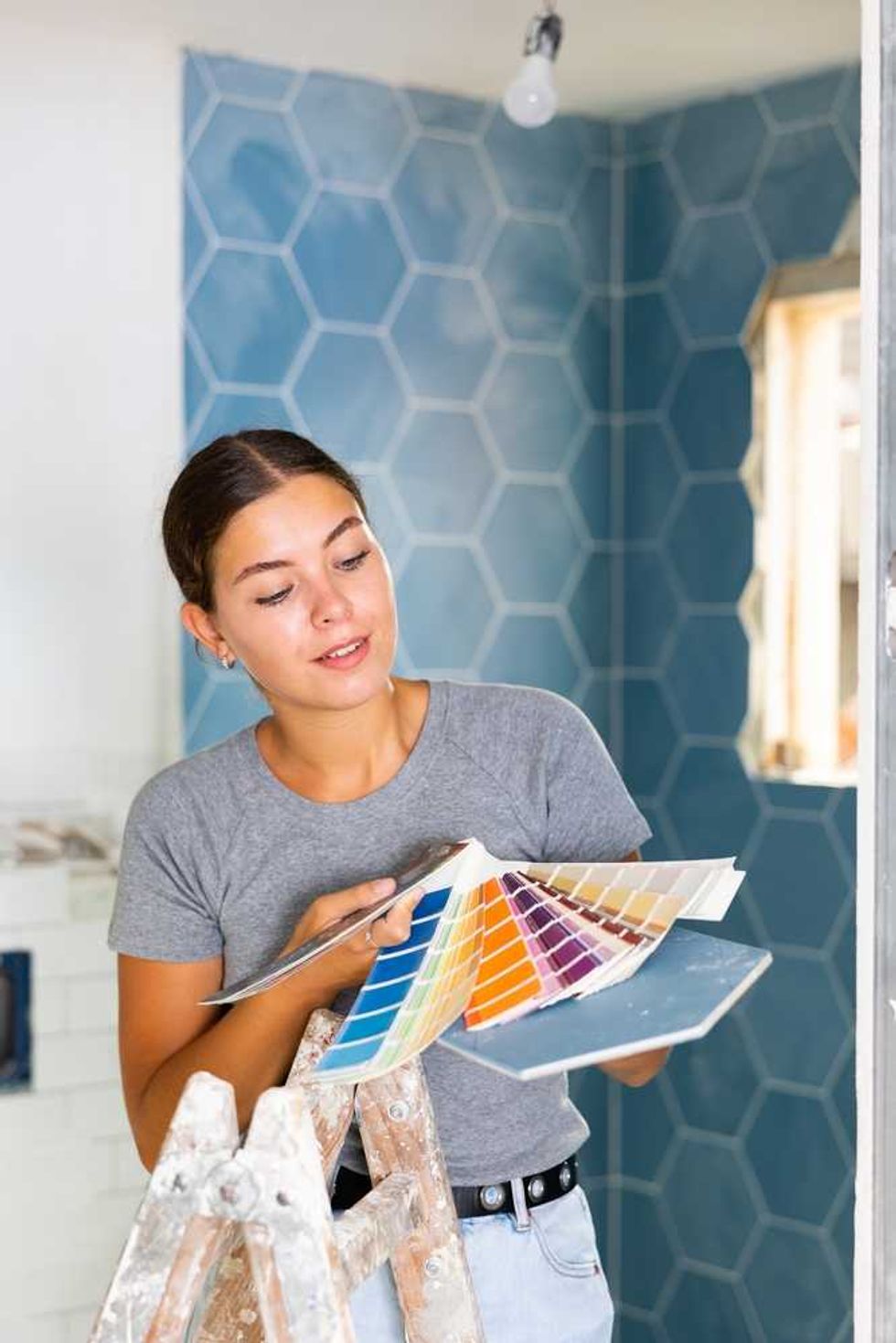 Woman on ladder choosing paint colors with hexagonal tiles in the background.