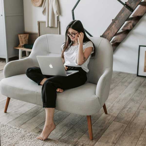 Woman on sofa using a laptop, wearing glasses and headphones in a cozy room.