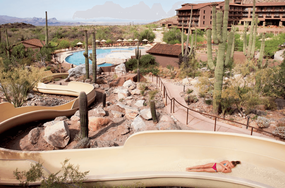 Woman on waterslide in desert resort, surrounded by cacti and rocks, leading to pool.