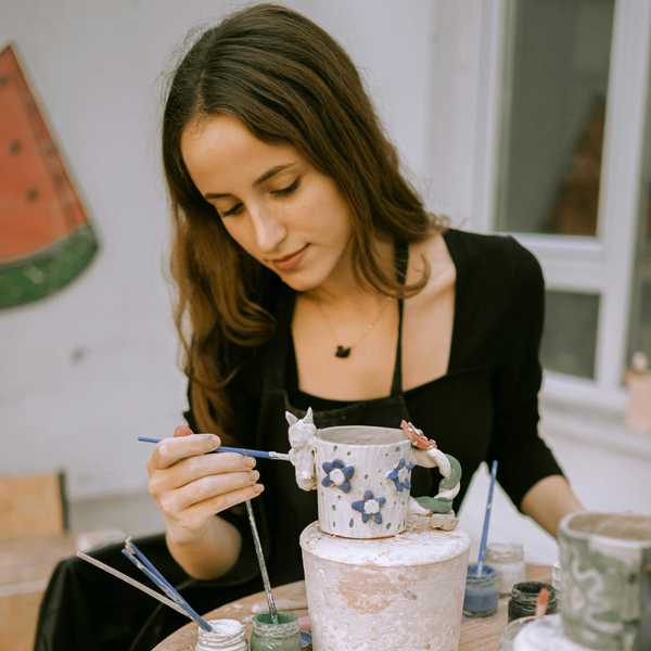 Woman painting a ceramic cup at a table with brushes and paints.